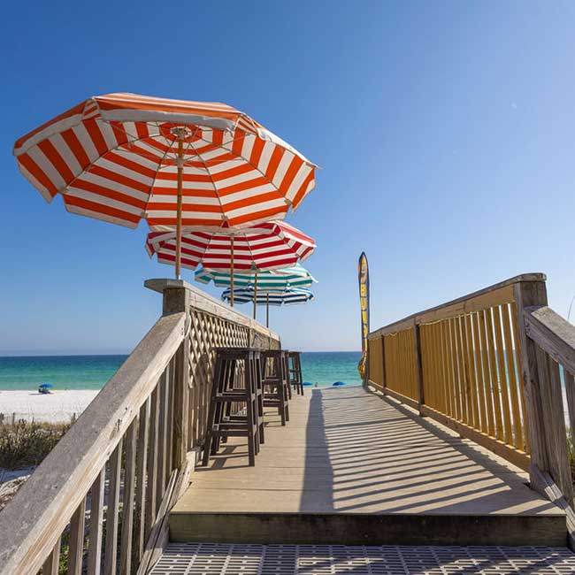 Wooden boardwalk to the beach at a beachfront resort with colorful umbrellas and chairs.