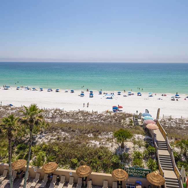 A sunny Fort Walton Beach scene with blue umbrellas, white sand, and palm-lined boardwalk.