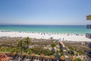 Aerial view from a penthouse of a sandy beach, blue umbrellas, and wooden boardwalk below.
