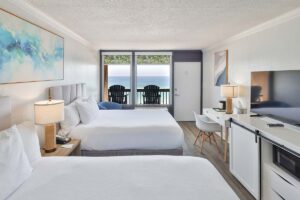 Modern penthouse hotel room with two beds, desk, TV, and balcony ocean view at a beachfront resort.