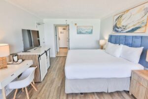 Modern beach hotel room with king bed, TV, desk, lamps, and minimalist decor.