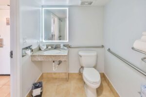 Accessible bathroom near hotel pool with toilet, grab bars, sink, mirror, towels, and tiled floor.