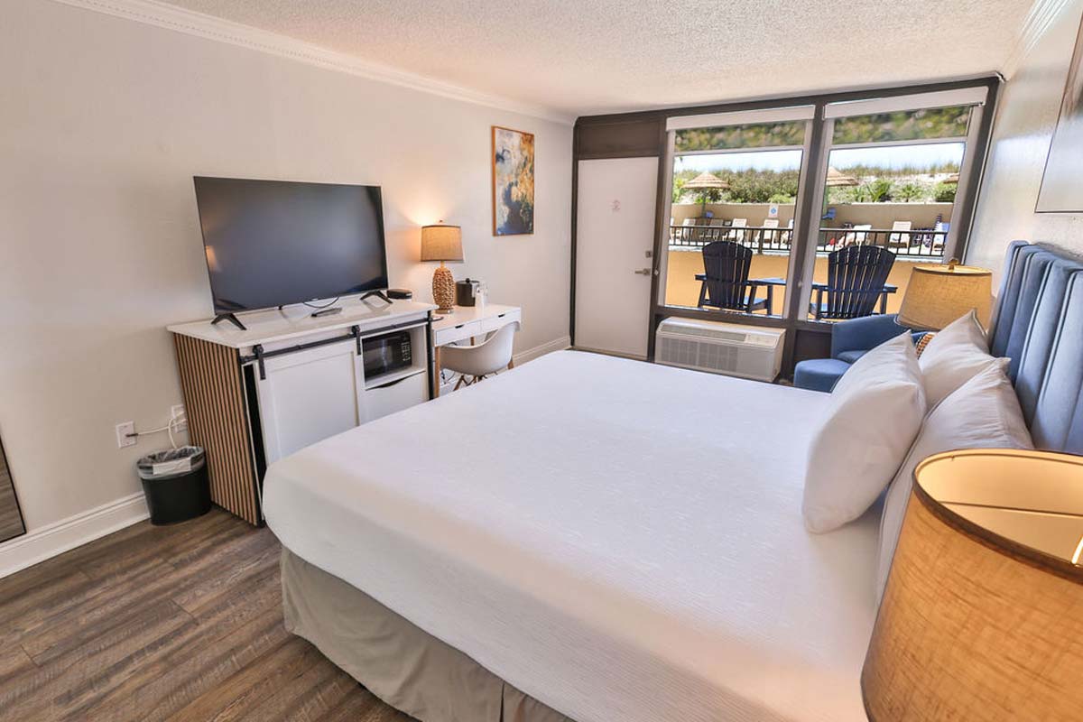 Penthouse hotel room with king bed, TV, desk, and balcony with chairs overlooking greenery.