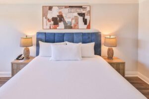 A neatly made hotel bed with blue headboard, two lamps, and abstract art above.