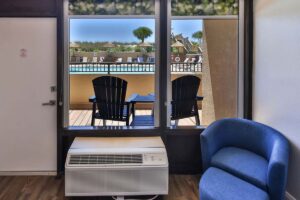 Blue chairs and hotel pool viewed through a window in a penthouse room with armchair and AC unit.