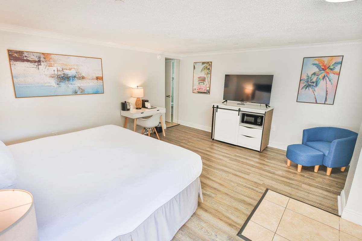 Bright Fort Walton Beach hotel room with king bed, TV, desk, blue chair, and palm art.