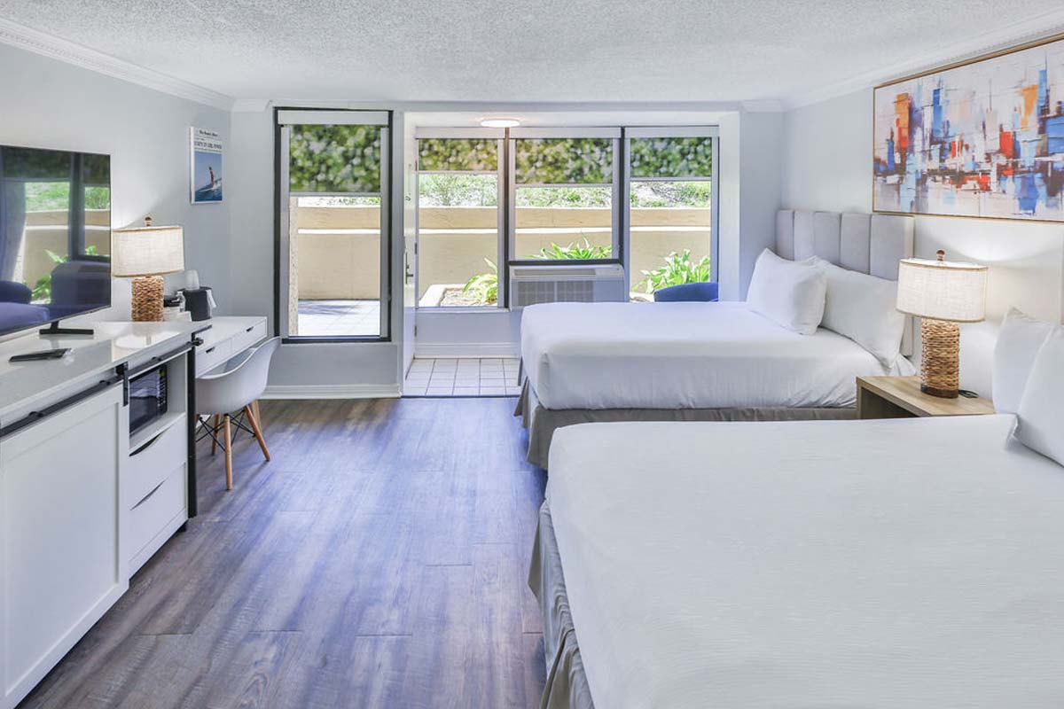 Modern hotel room with two beds, desk, TV, and views of the hotel pool amid lush greenery.
