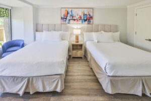 Hotel room with two queen beds, nightstand, lamp, blue chair, and abstract hotel wall art.