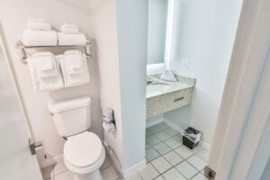 Small, clean hotel bathroom with a toilet, towels on a rack, sink, mirror, and trash bin.