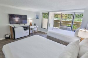 Bright Fort Walton Beach hotel room with two beds, desk, TV, mini fridge, and lush window view.