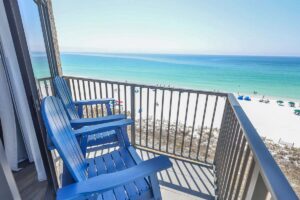 Two blue chairs on a penthouse balcony overlook a sandy beach and turquoise ocean on a sunny day.