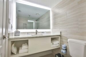 Modern bathroom with hotel pool vibes, large illuminated mirror, sink, towels, and toilet on right.